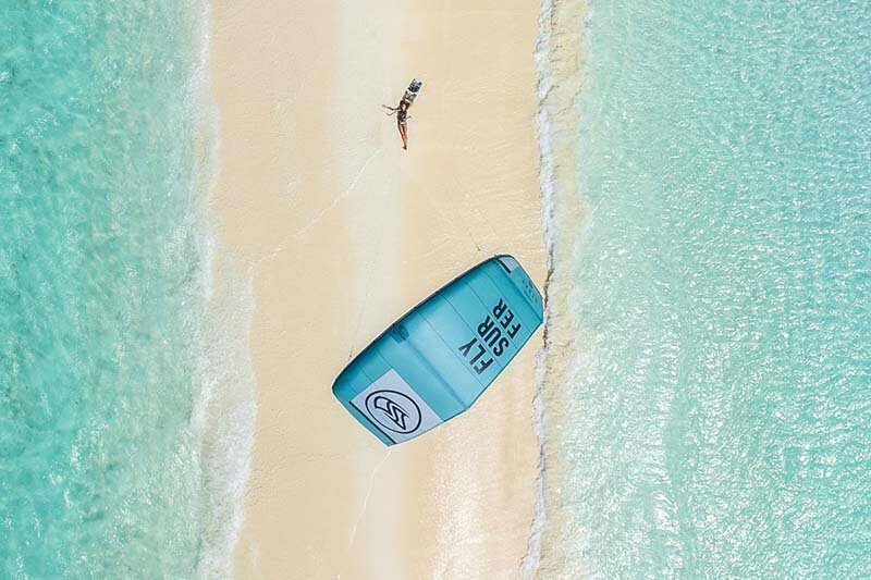 Flysurfer Kite - Frau liegt auf dem Rücken am Strand. Links und rechts Wasser und der Kite schwebt über ihr. Das Foto ist weit über dem Kite mit Sicht nach unten geschossen