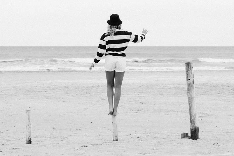 Mystic Boarding - Frau steht auf einem Holzpflock am Strand. Das Wasser ist im Hintergrund zu sehen