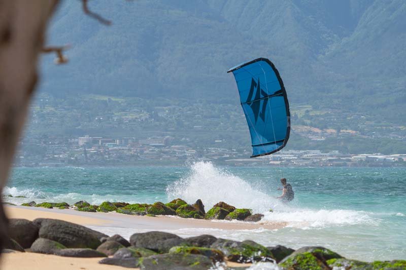 Naish Triad Kite - Kiter fährt von Brandung am Strand in Richtung Meer. Im Hintergrund ist eine Große Bergformation zu sehen und am Strand liegen große, mit Algen bedeckte Steine