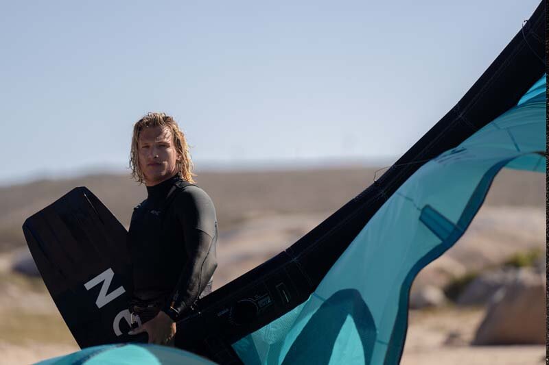 North Kiteboarding - Kitesurfer in Neoprenanzug mit Board unter dem Arm und Kite in der Hand steht am Strand in Nahaufnahme