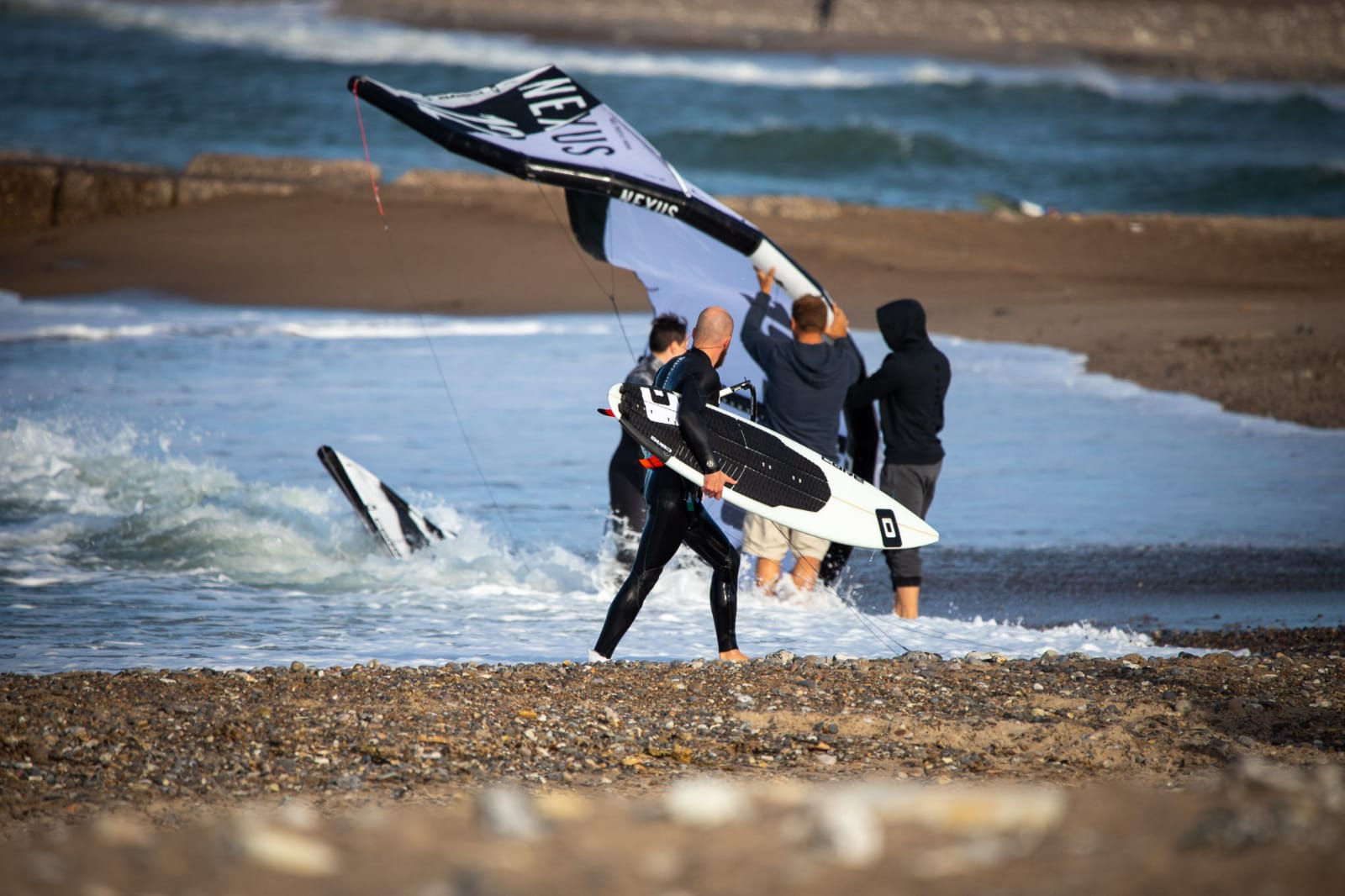Auf dem Bild erkennen wir einen Kitesurfer der mit seinem Waveboard aus dem wasser geht, der Kiteschirm wird von drei Menschen festgehalten.