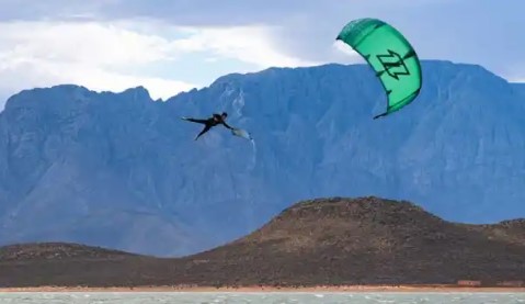 zu erkennen ist ein Kitesurfer der in Schwindelliger Höhe einen Kiteloop-boardoff springt. Hinter ihm sieht man Hohe Berge.