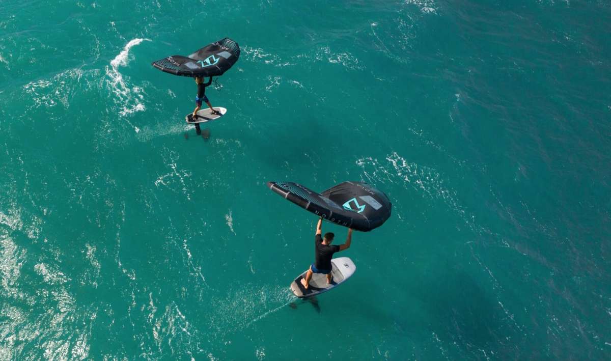 Wir sehen zwei Wingsurfer mit Foil die durch klarstes türkises Wasser fahren.